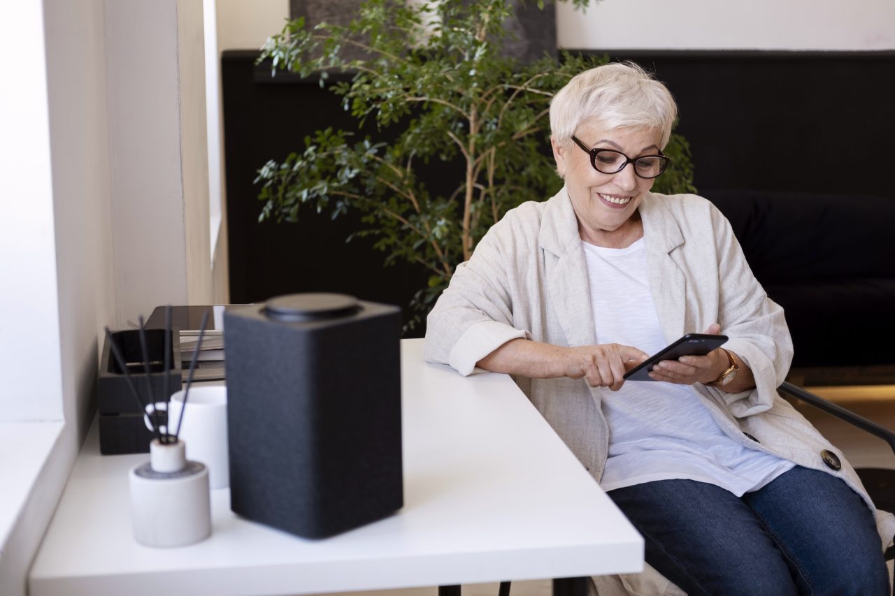 Idosa mexendo no celular sorrindo, ela está em casa e usa camiseta branca, calça jeans e casaquinho de linho. Na frente tem uma pequena caixa de som.