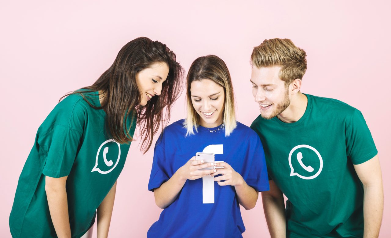 Na imagem com fundo rosa claro tem um homem e uma mulher usando blusa com logo do WhatsApp e no meio tem uma mulher com blusa com logo do facebook mexendo no celular sorrindo.