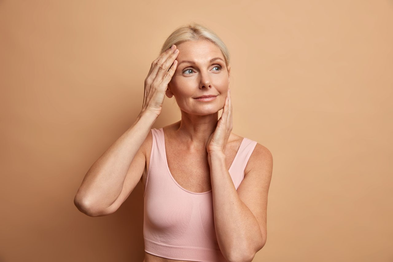 Senhora de cabelos grisalhos e usando top rosa, está com as mãos tocando levemente o rosto, pois ela fez algum tratamento facial.