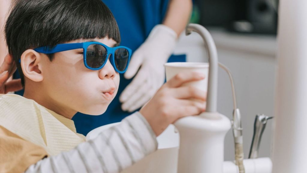Criança no dentista fazendo bochecho perto da pia enquanto segura copo com o enxaguante.