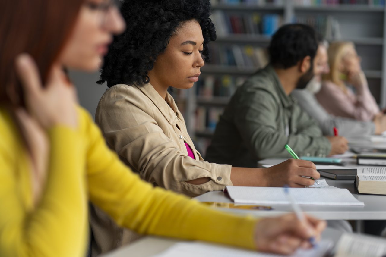Vários adultos em uma biblioteca estudando com caderno e livros.