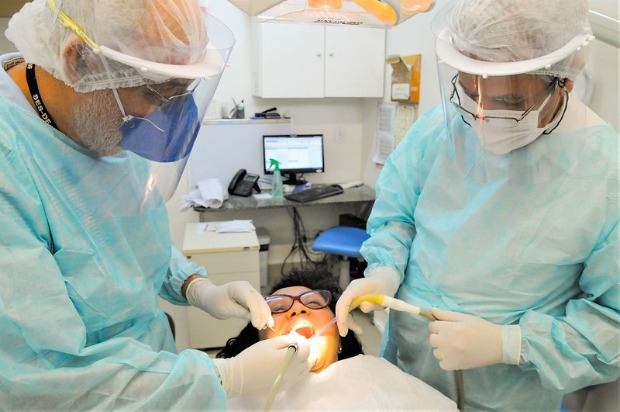 Duas dentistas estão com todos os EPIs atendendo paciente no consultório.