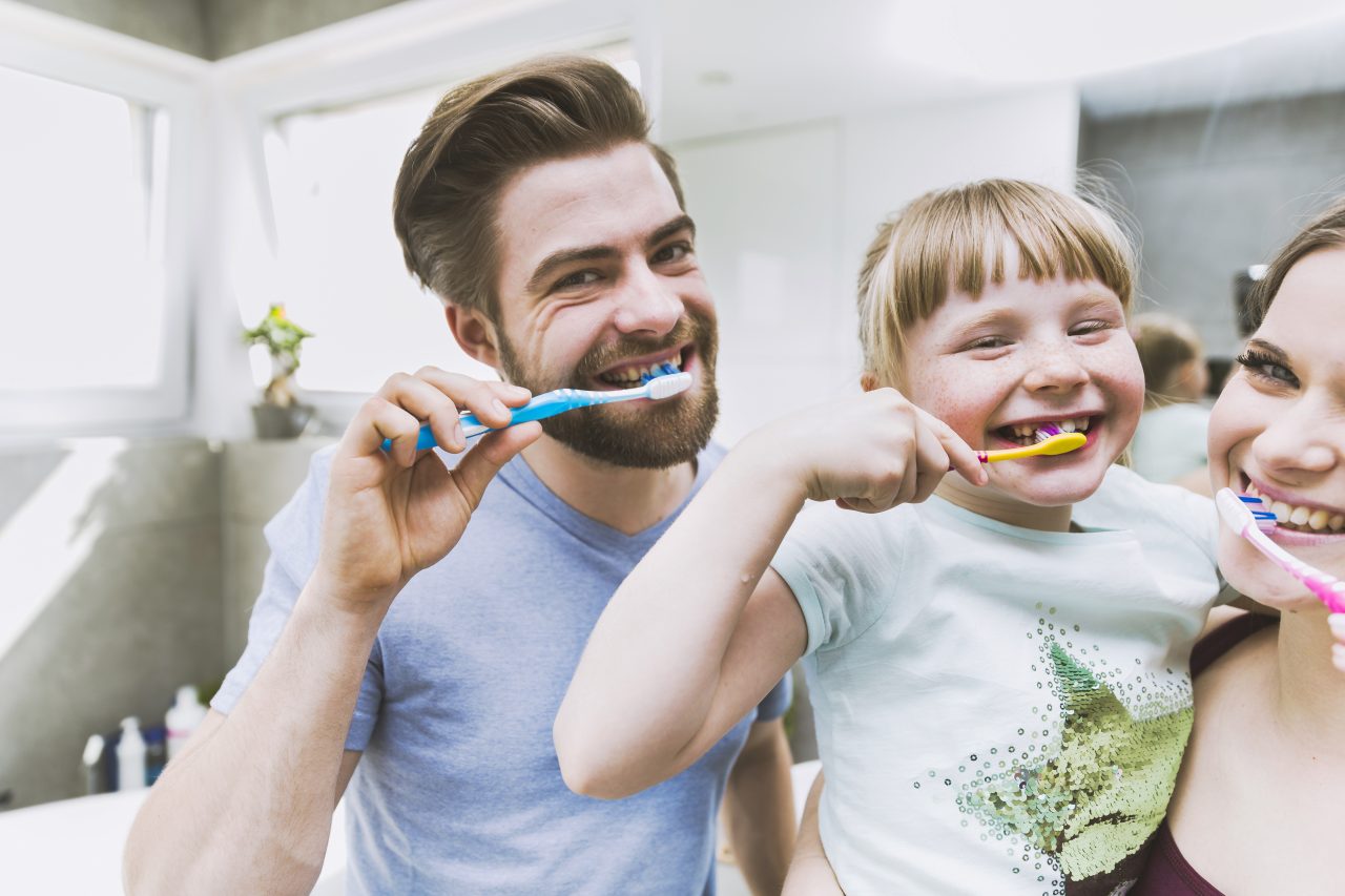 Menina escovando os dentes no colo da mãe e com o pai ao lado, todos estão no banheiro felizes;