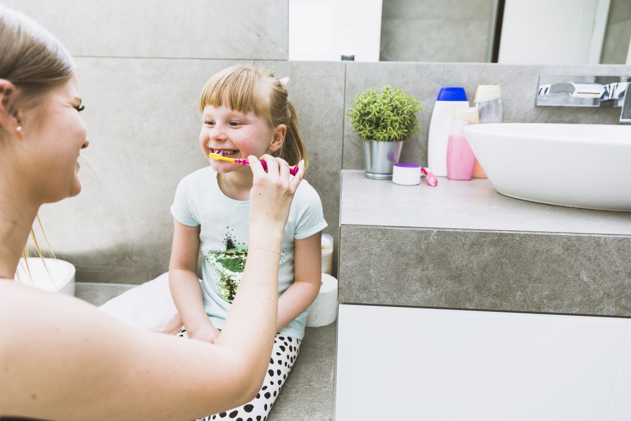 Mãe escovando os dentes da filha que está sentada na bancada do banheiro.