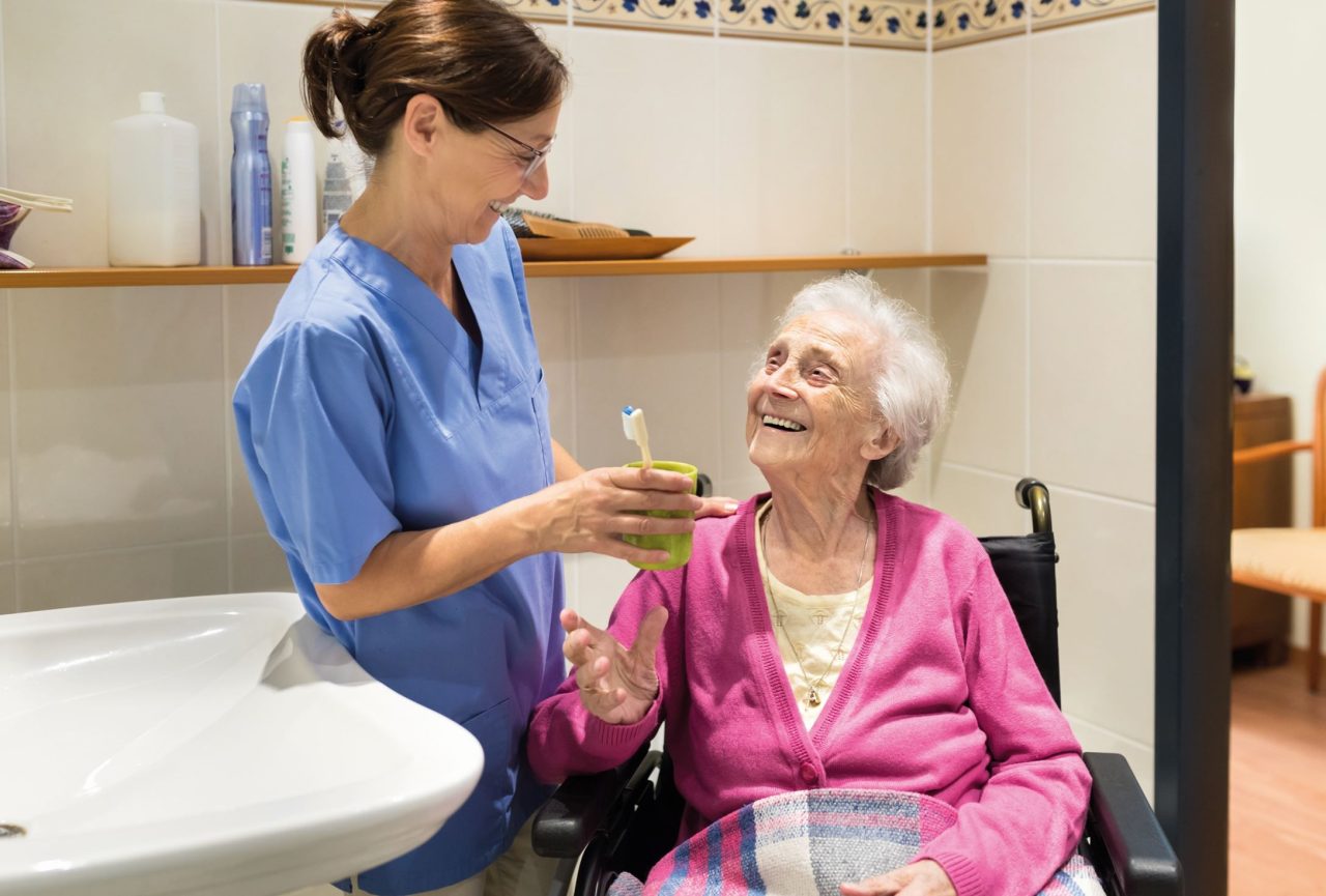 No banheiro, dentista está segurando xícara com escova de dente dentro e entregando para senhora idosa em cadeira de rodas