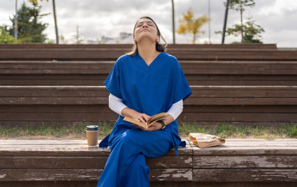 Mulher com uniforme médico está sentada em banco de praça sozinha com livro, saco de papel com comida e copo de café.