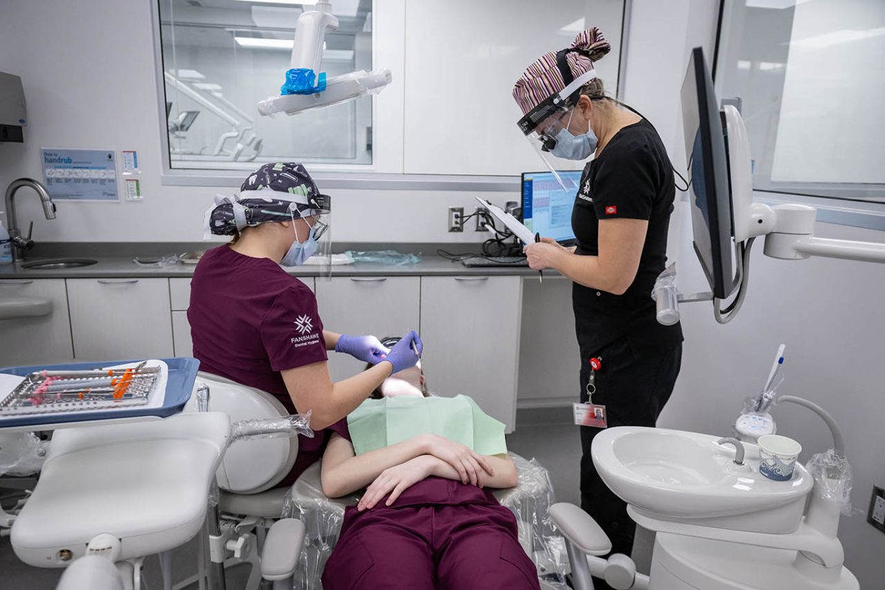 Dentista atendendo paciente, e técnica em saúde bucal em pé ao lado com prancheta.