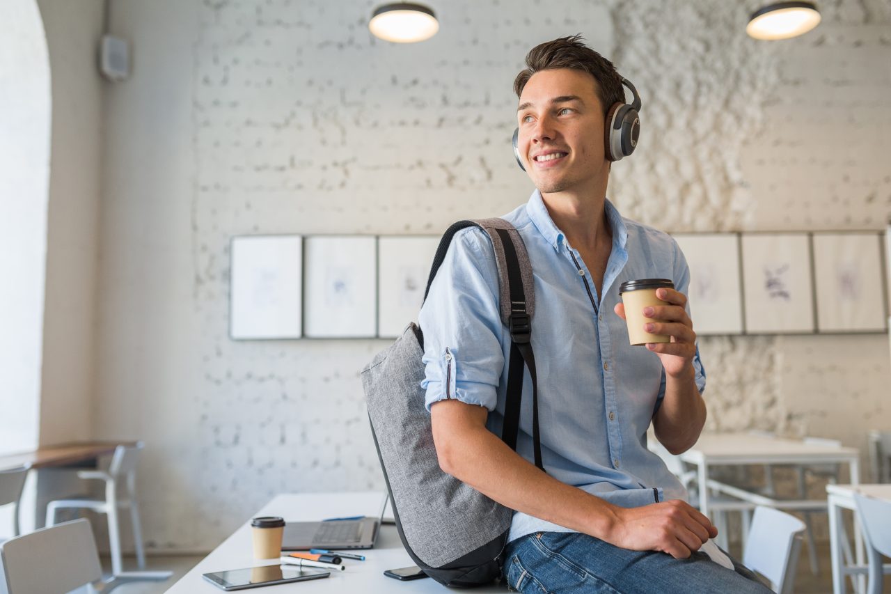 Home jovem está sentado sob uma mesa sorrindo. Ele usa mochila, fones de ouvido e seguro um copo descartável de café.