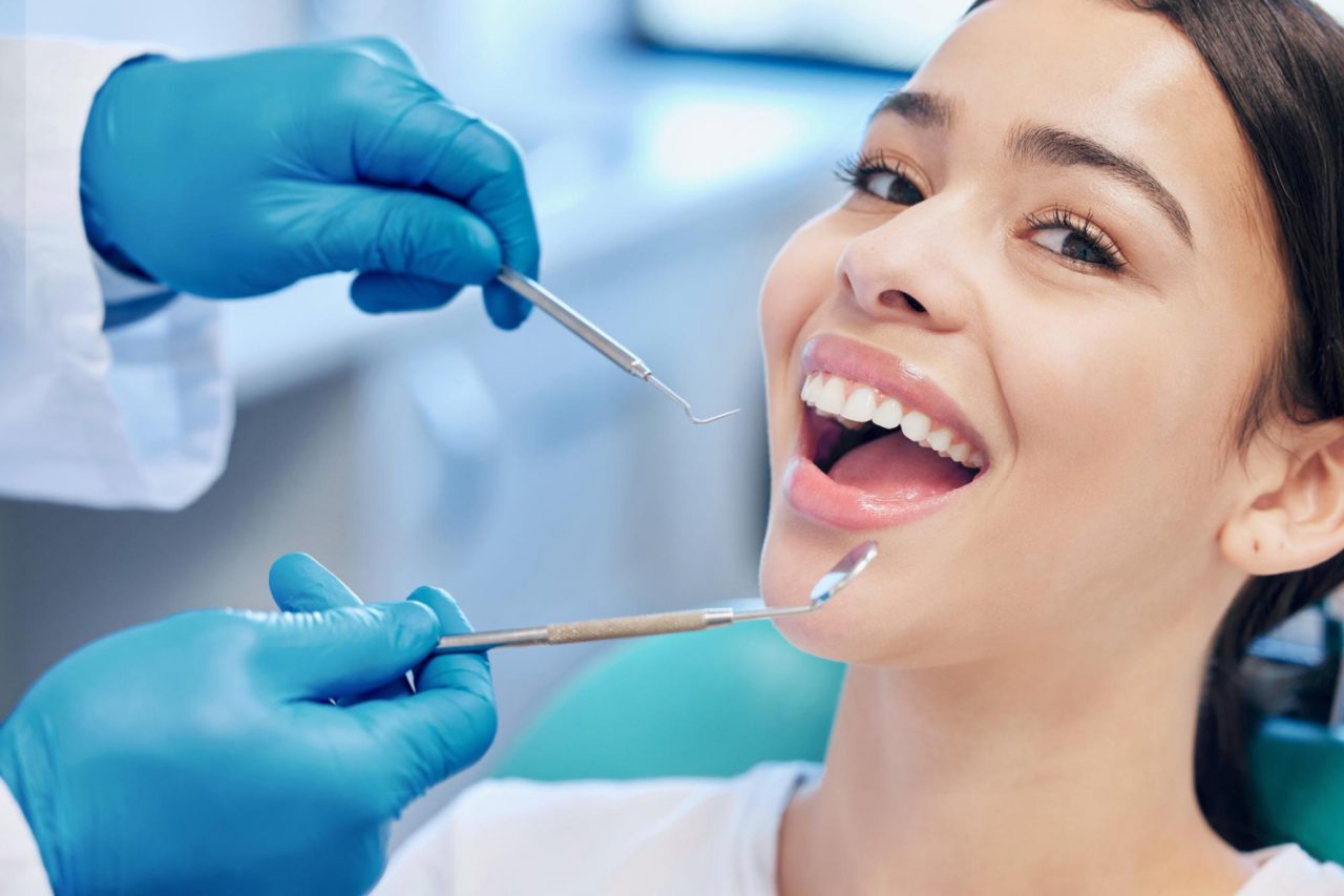 Paciente em consultório está olhando para a câmera sorrindo de boca aberta, enquanto dentista se aproxima com instrumentos odontológicos para avaliação.