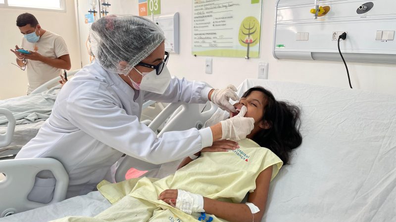 Na odontologia hospitalar, a dentista está olhando os dentes de uma garotinha que está na cama do hospital acordada.