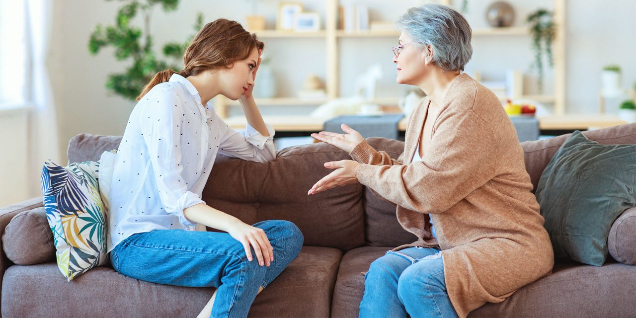 Mãe idosa aconselhando a filha que a olha com olhar de tédio. Ambas estão sentadas no sofá de casa.