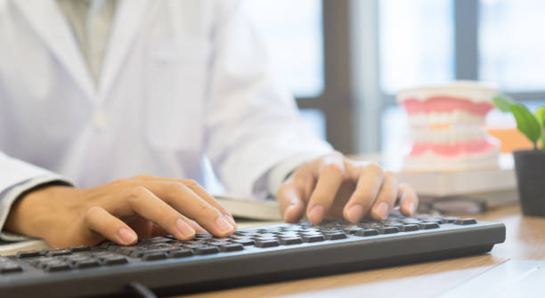 dentista digitando em computador, ao lado tem um vasinho de plantas e um manequim odontológico.