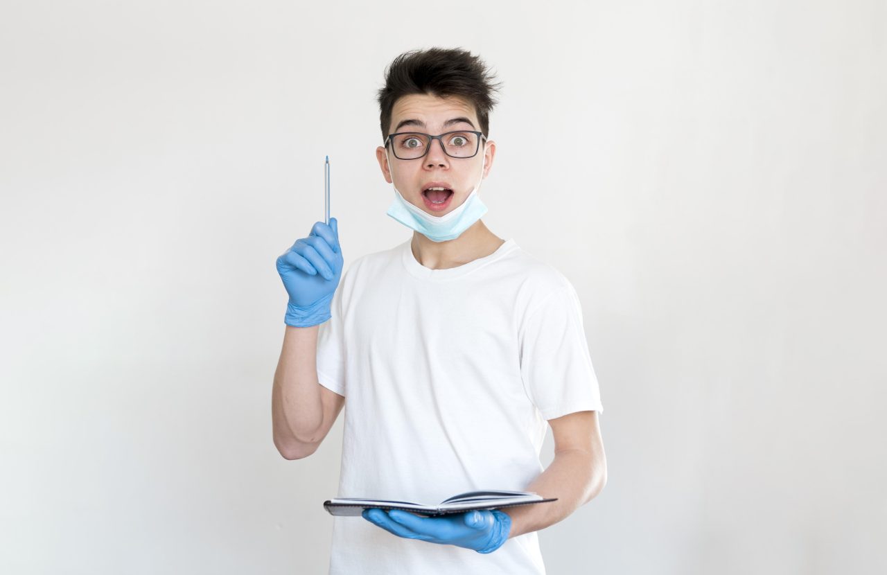 Jovem garoto de luvas e máscara descartável está com caderno aberto na mão e caneta apontando para cima com a outra como se tivesse tido uma ideia.