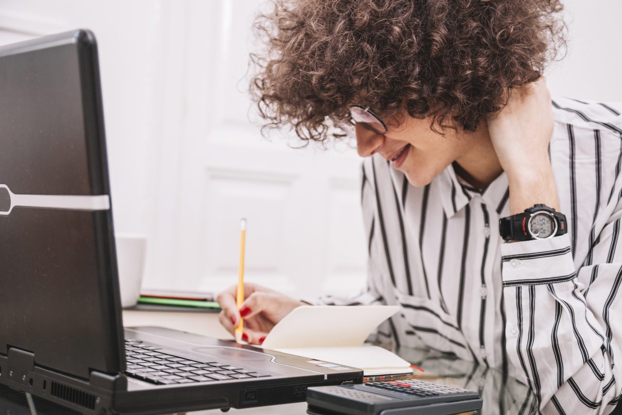 Mulher com notebook na mesa, está anotando algo em seu caderno.