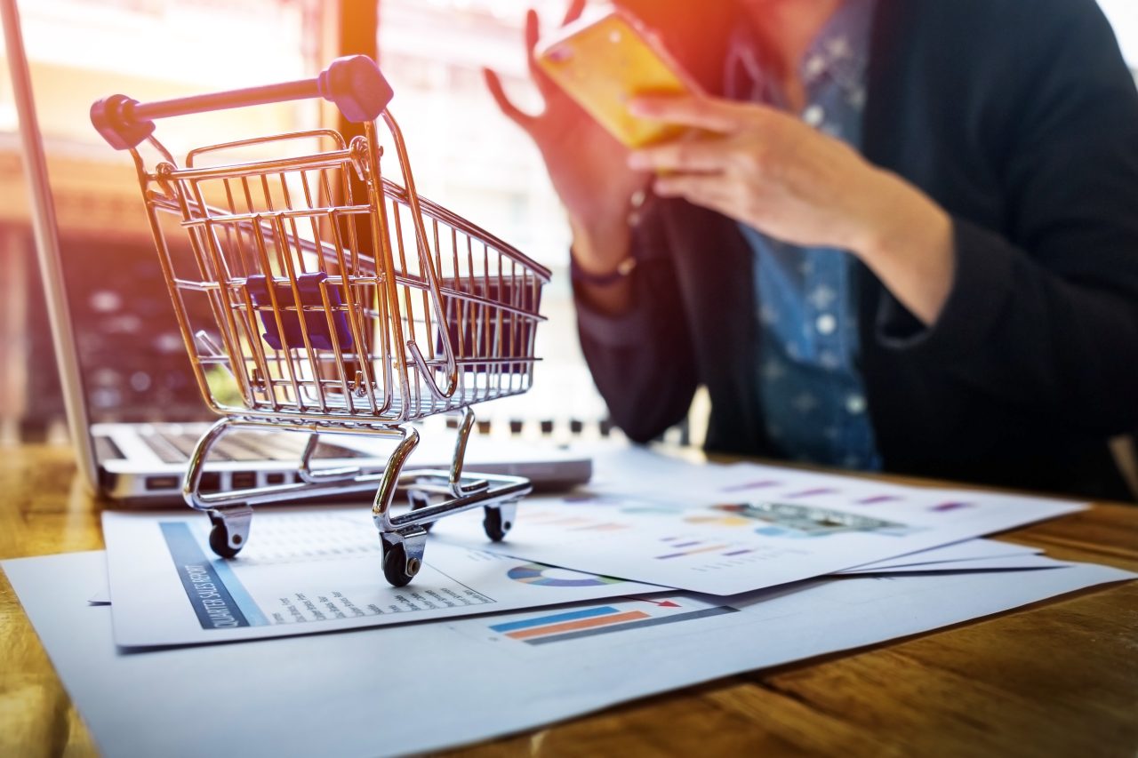 Mulher em escritório fazendo compra em celular, em sua frente na mesa tem um notebook, um carrinho de compras pequeno, e papéis espalhados.
