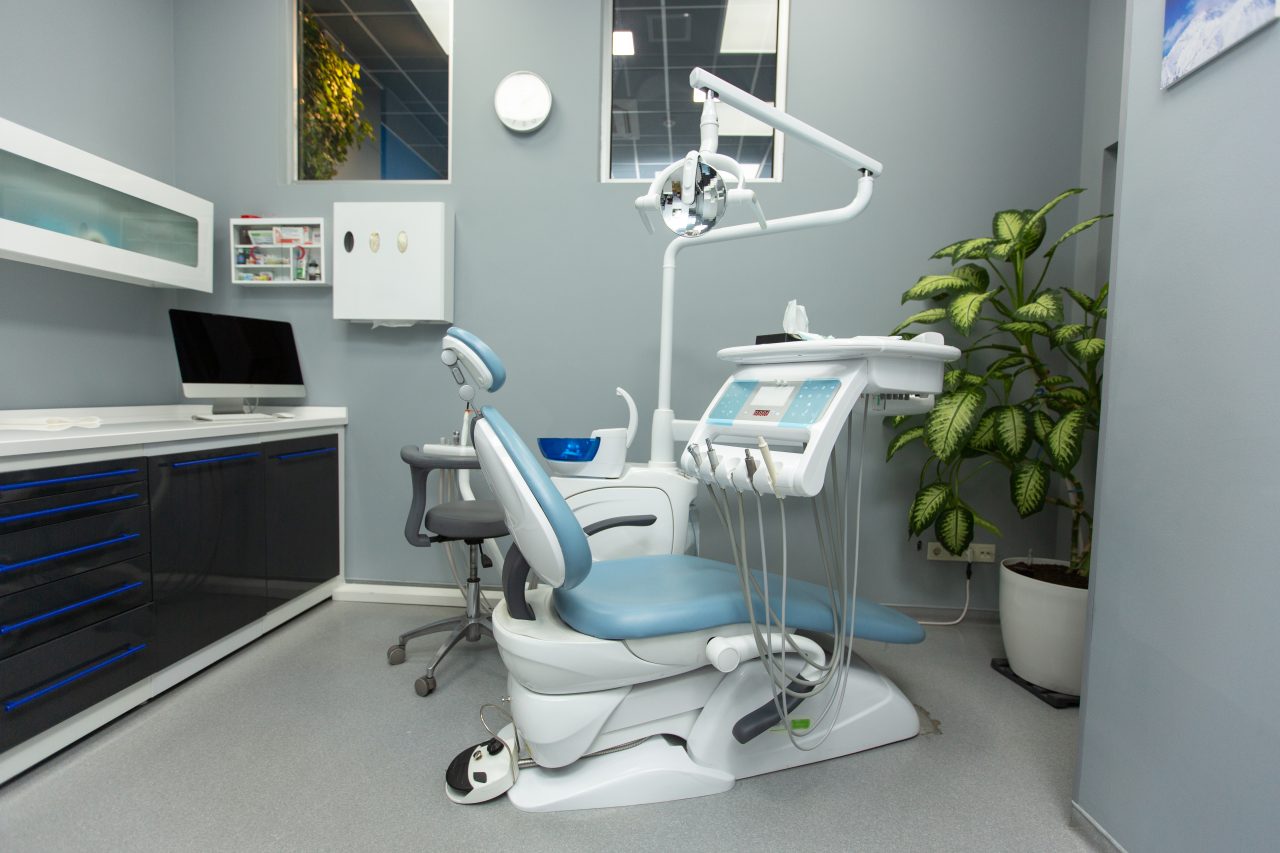 Consultório de dentista com cadeira odontológica, bancada preta com computador, e vaso de plantas.