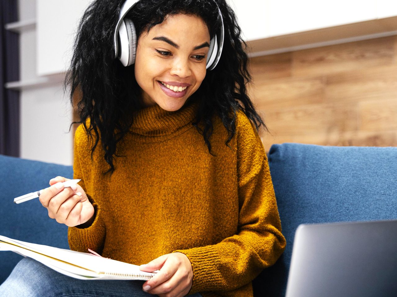 Jovem mulher está estudando no computador com fones de ouvido, sentada em seu sofá.