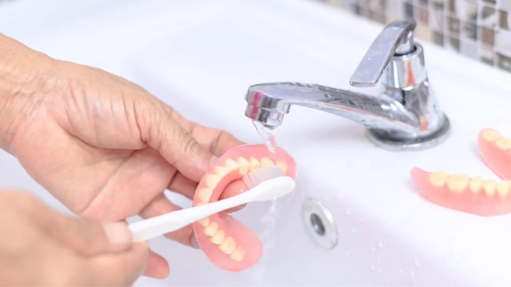 Limpando prótese dentária removível com escova de dente na pia do banheiro.