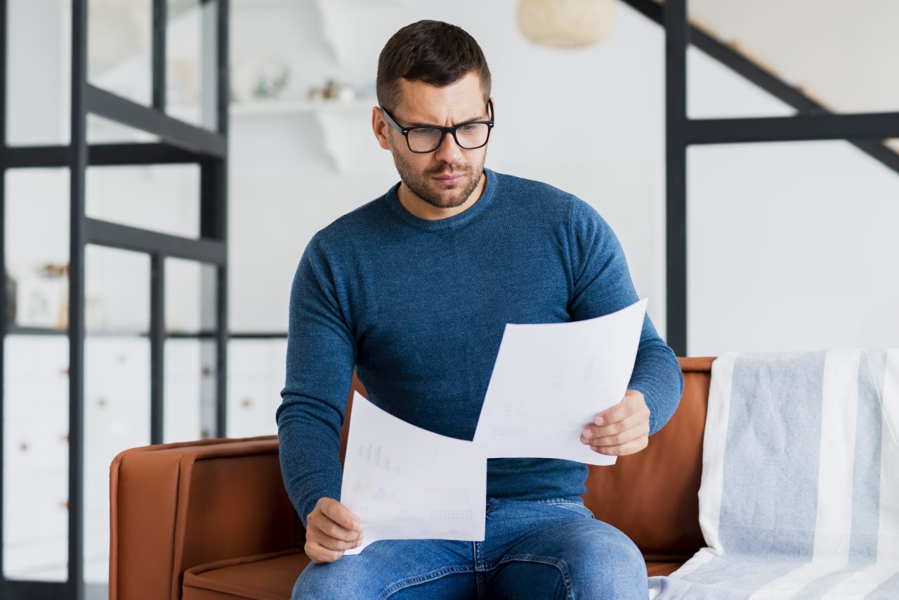 Homem lendo contrato com rosto sério no sofá de sua casa.
