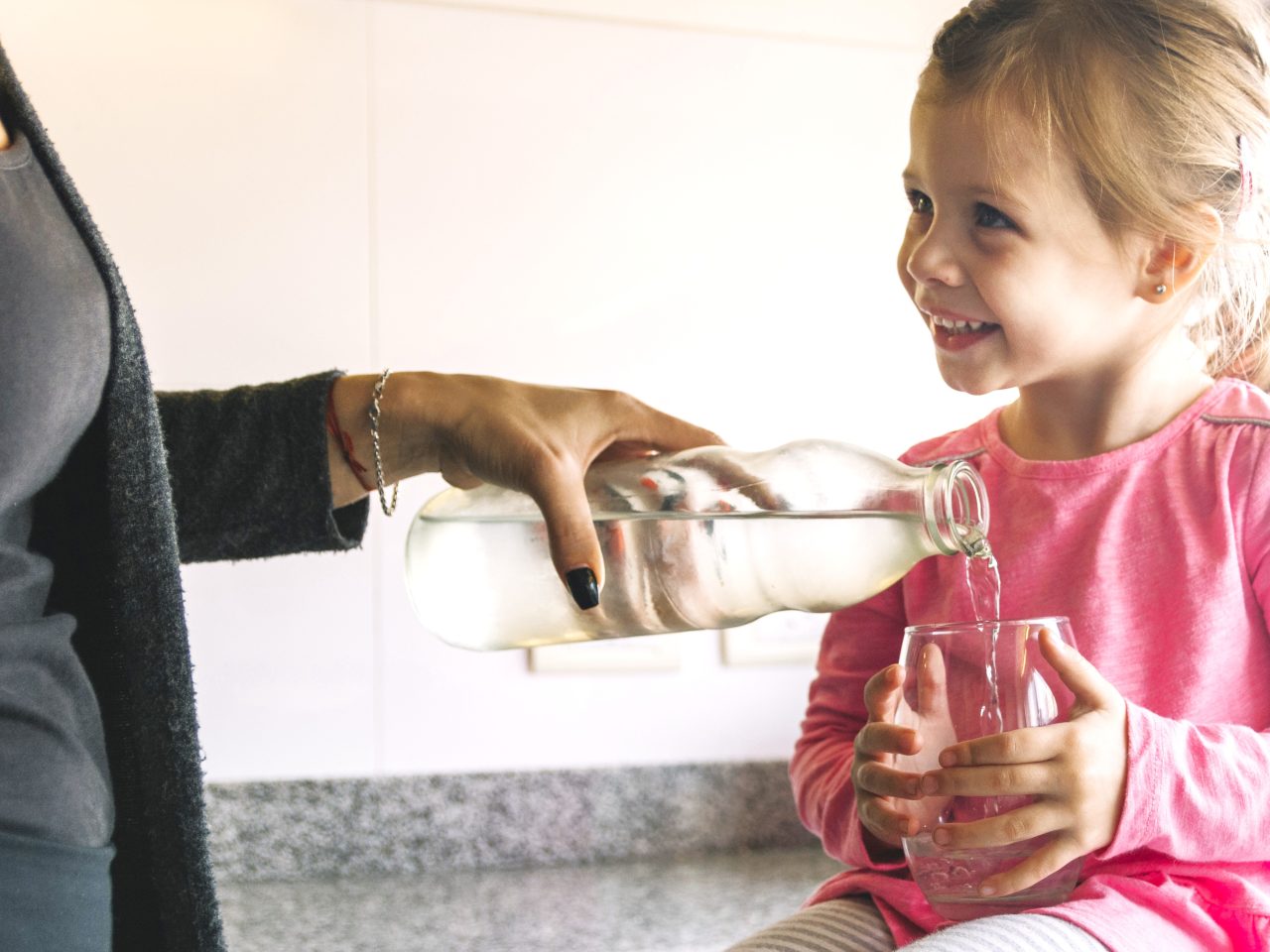 Garotinha sentada em balcão da cozinha segurando um copo transparente, enquanto sua mãe enche com água.