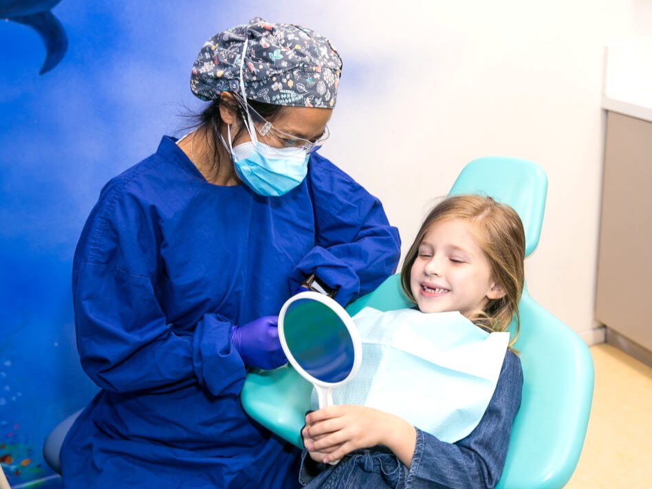Garotinha em consultório odontológico vendo seus dentes no espelho, com a dentista ao lado.