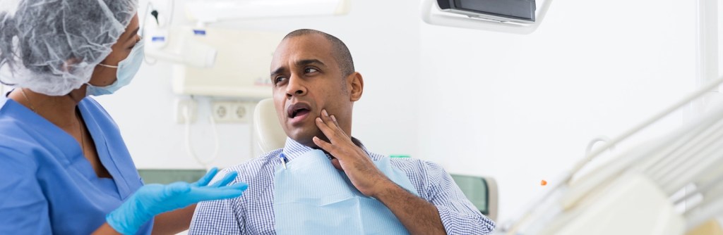 Paciente está com dor de dente apertando a bochecha, e a dentista está com postura de dúvida.