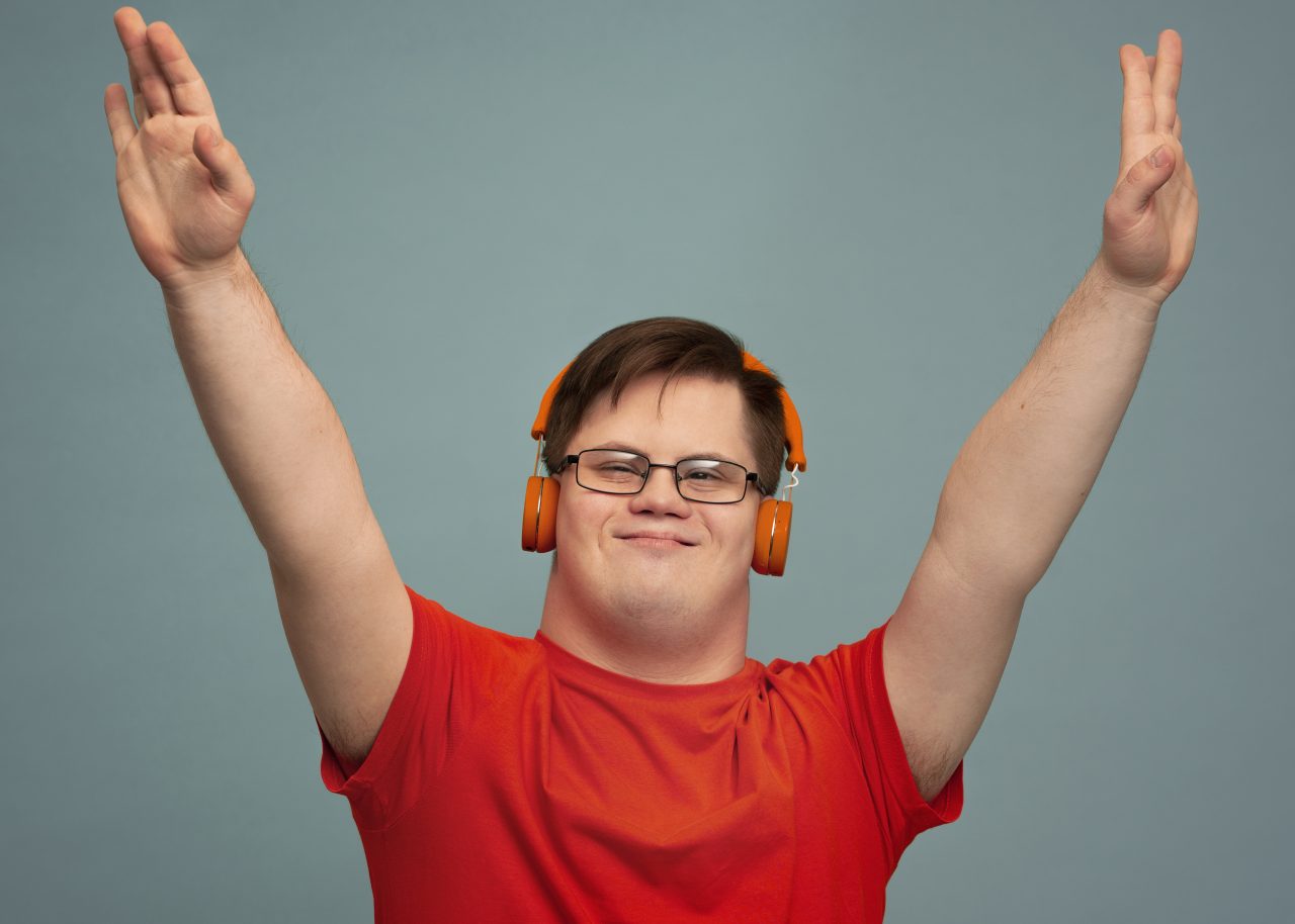 Rapaz com síndrome de down está de fones de ouvido e com os braços para cima feliz.