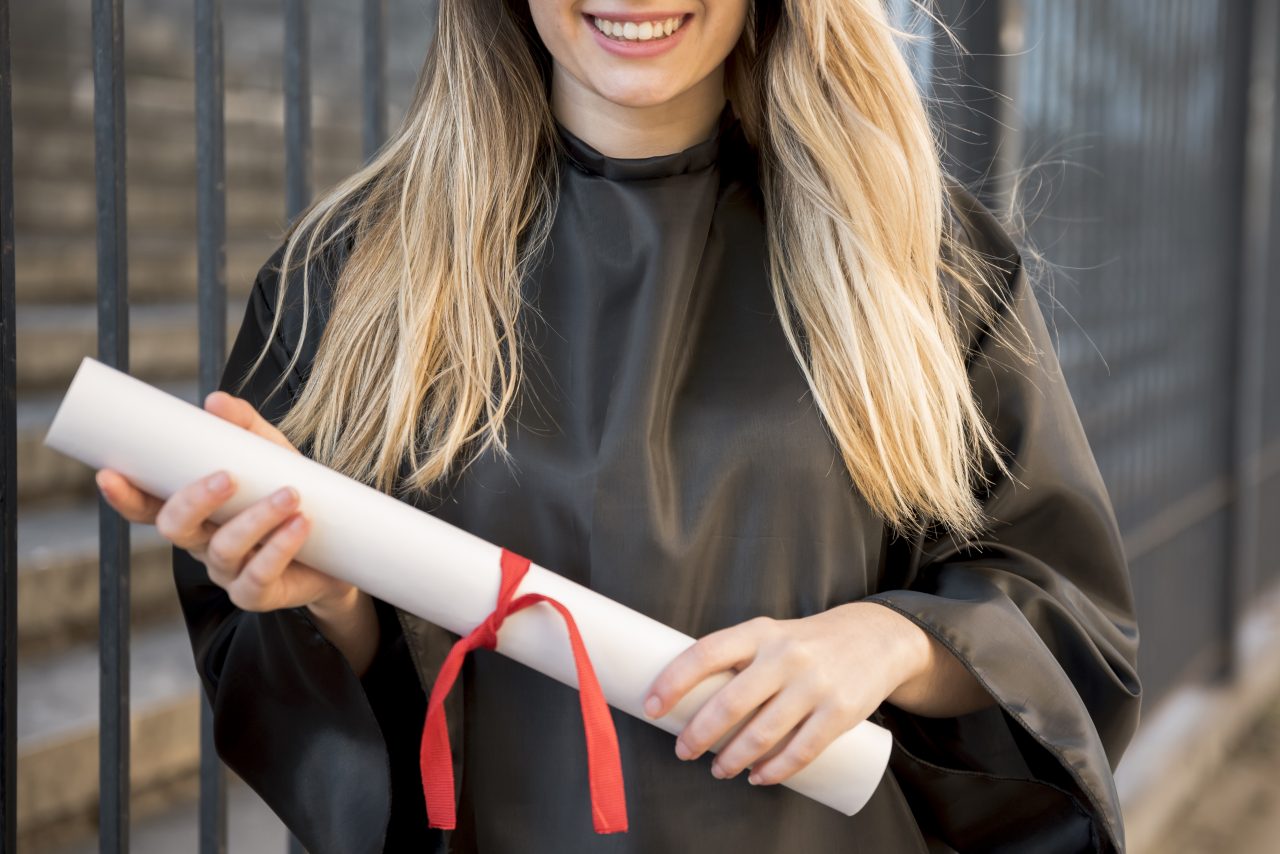 Jovem mulher de cabelos loiros está no seu dia de formatura e segura diploma na mão.