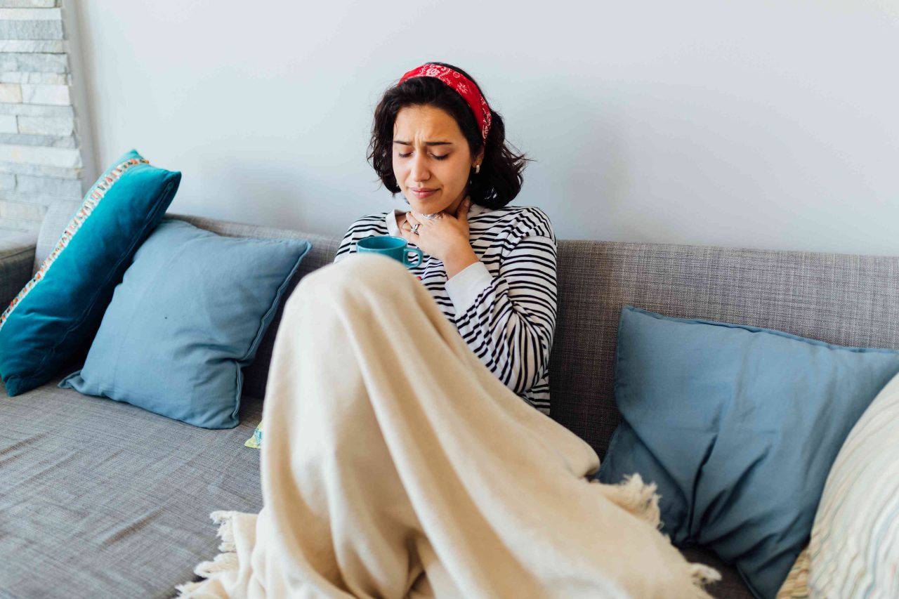 Mulher está sentada no sofá com coberta, segurando xícara e com a outra mão apertando a garganta que está com dor.