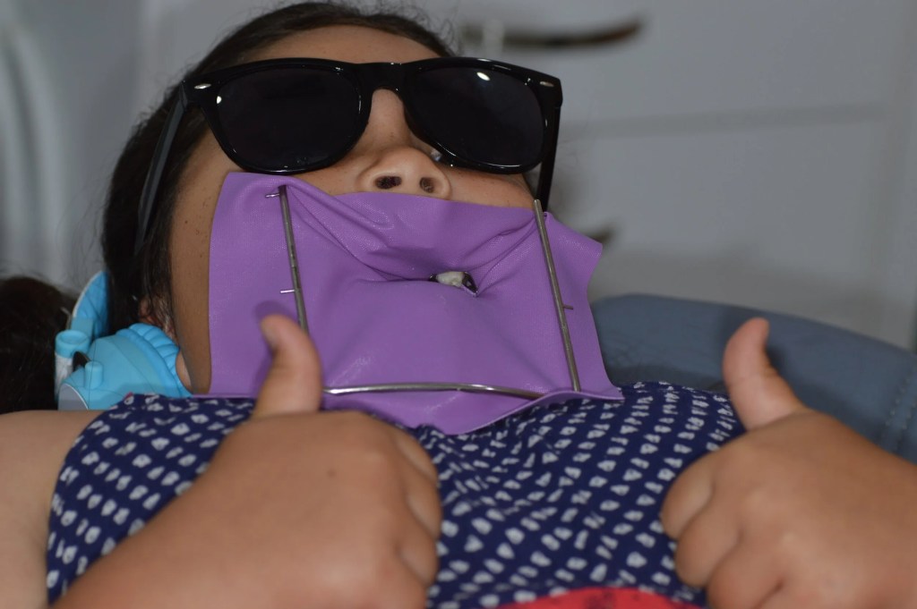 Imagem de criança com isolamento absoluto segurado por grampos odontológicos está fazendo sinal de joinha com as duas mãos.