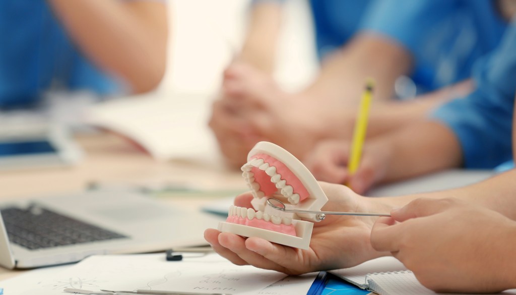 Dentistas ao redor de mesa, o fundo está desfocado, sendo focado um deles que está treinando com manequim odontológico na mão com espelhinho.