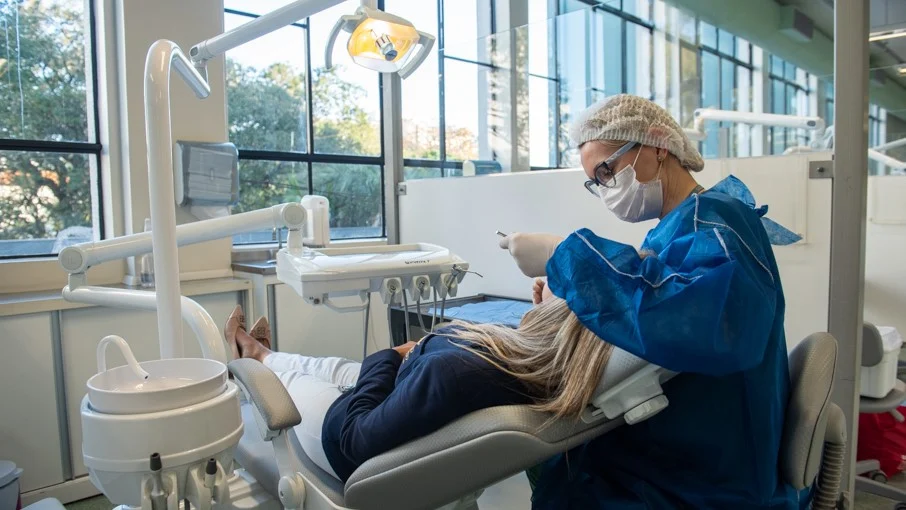 Dentista atendendo paciente em clínica com divisórias para separar, e as janelas são de vidro trazendo luz natural.