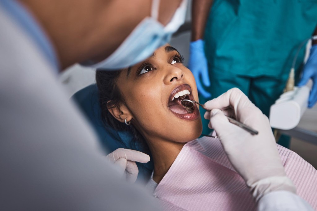 Dentista analisando com espelho clínico o interior da boca da paciente. No fundo o assistente.