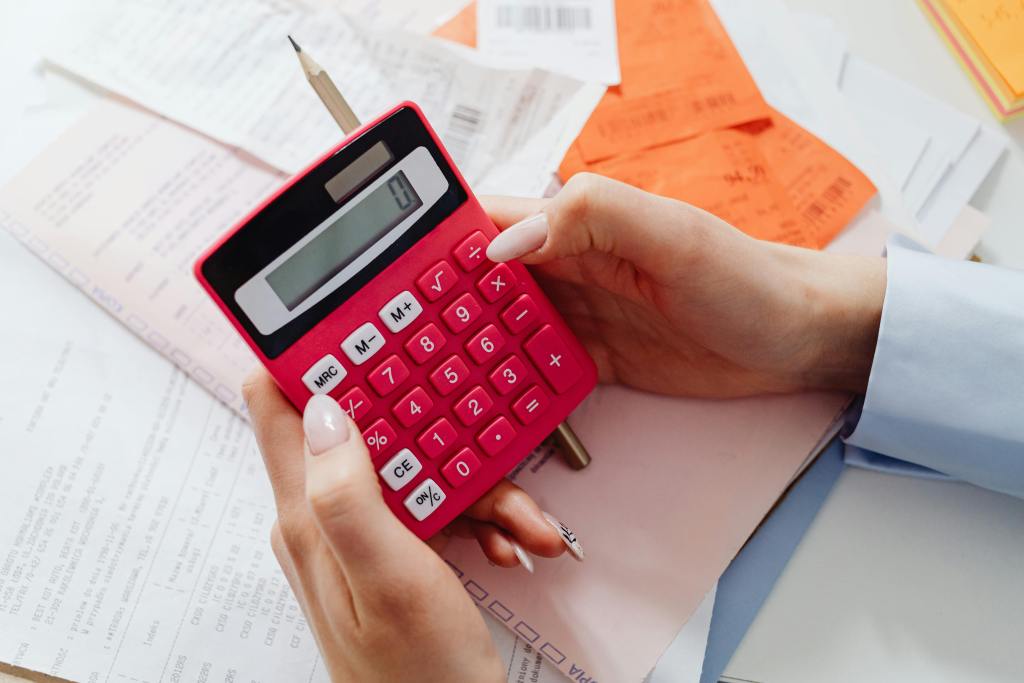 Mulher vai clicar em dividir na calculadora vermelha. Tem vários papéis com informações na mesa. Assim, ela pode calcular o ticket médio da clínica.