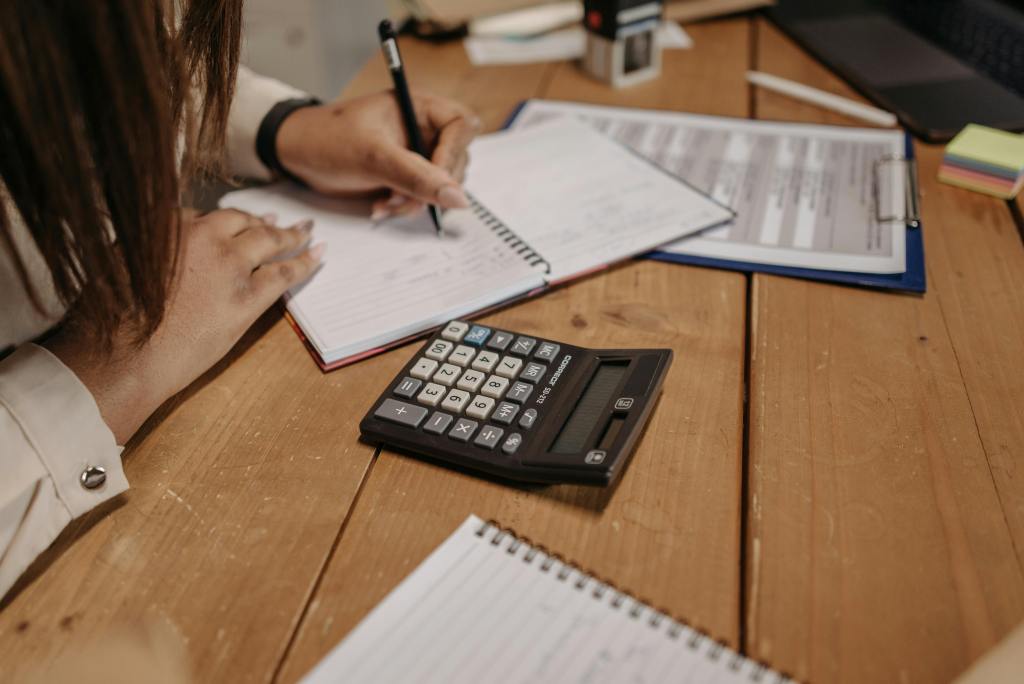 Mulher escrevendo em caderno com calculadora do lado.